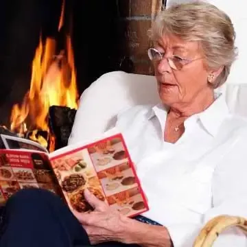 Eldre dame leser ved peis